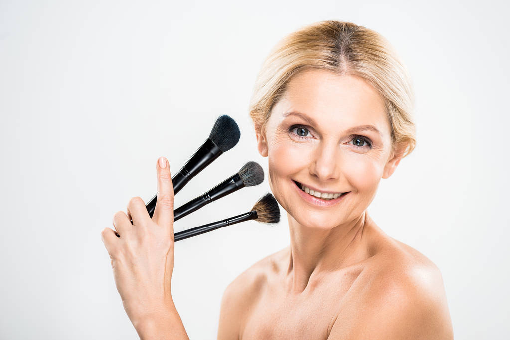 beautiful and blonde mature woman looking at camera and holding cosmetic brushes on grey background  - Photo, Image