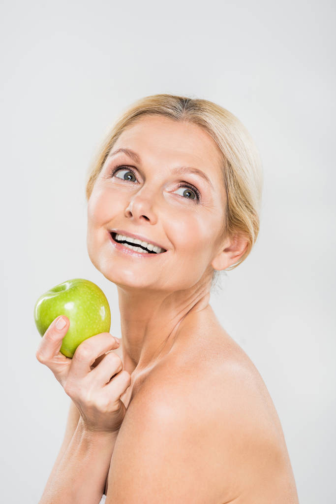 beautiful and smiling mature woman holding green ripe apple and looking away isolated on grey - Photo, Image