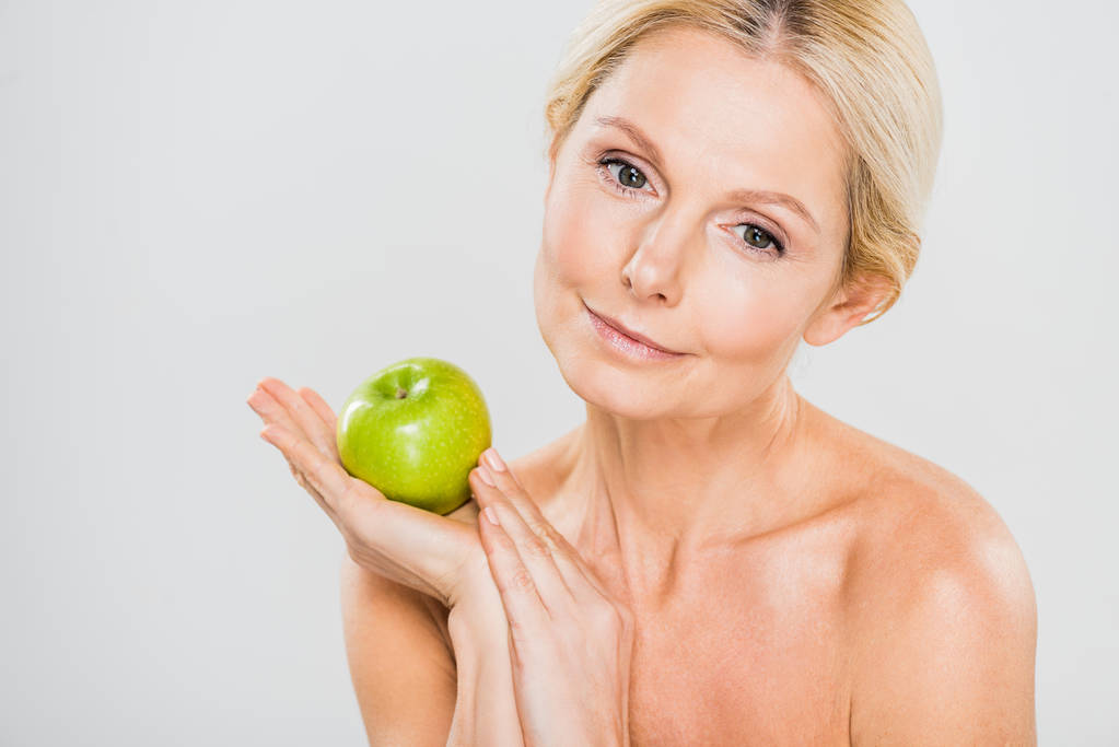красивая и зрелая женщина держит зеленое спелое яблоко и смотрит в сторону на сером фоне
  - Фото, изображение