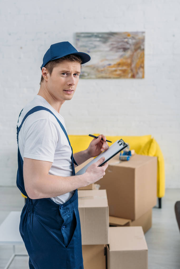 handsome mover holding clipboard and looking at camera near cardboard boxes in apartment - Photo, Image