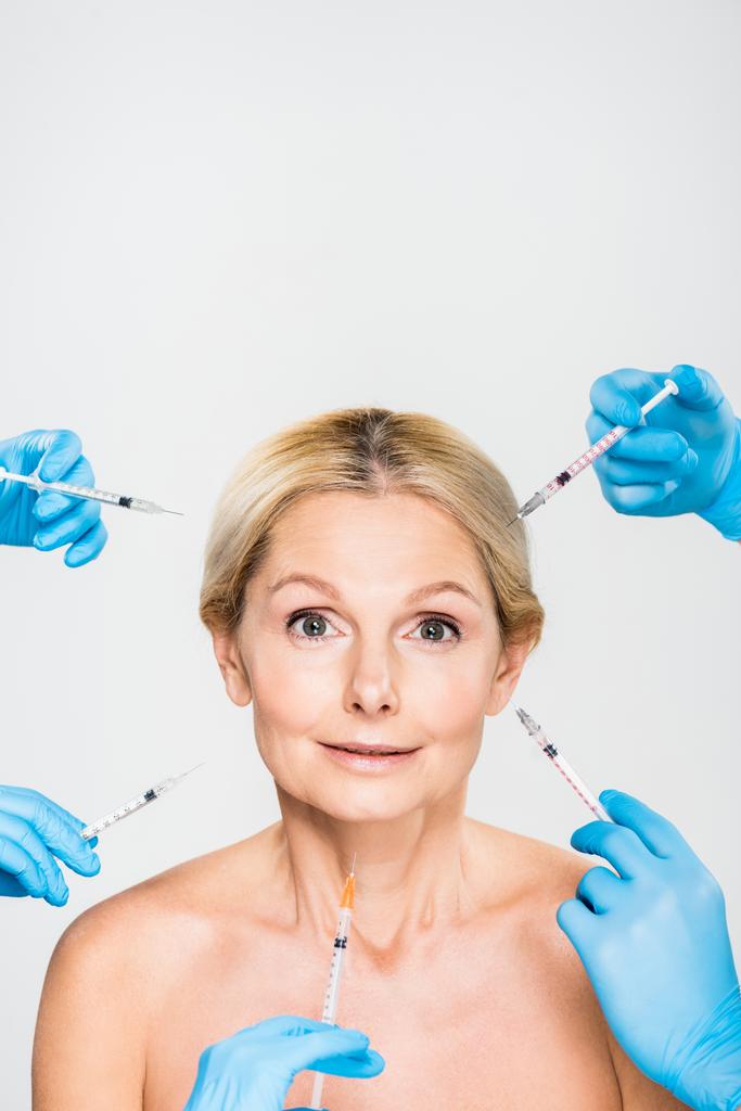 beautiful and mature woman looking at camera with cosmetologists holding syringes - Photo, Image