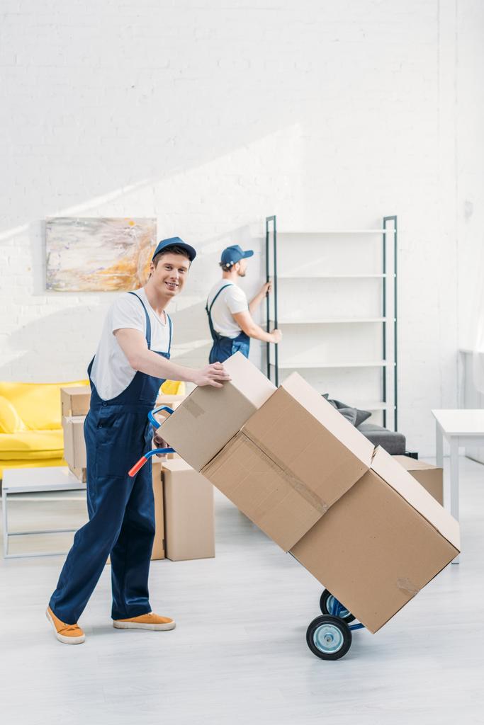 два перевозчика в униформе, перевозящих картонные коробки и мебель в квартире
 - Фото, изображение