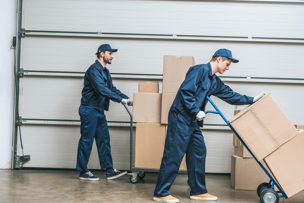 two movers in uniform using hand trucks while transporting cardboard boxes in warehouse - Photo, Image