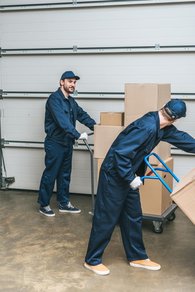 dois movers em uniforme usando caminhões de mão durante o transporte de caixas de papelão no armazém
 - Foto, Imagem