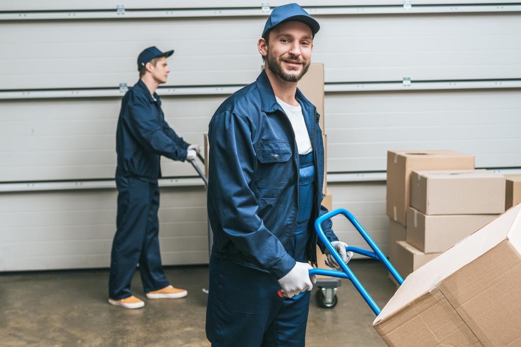 two movers in uniform transporting cardboard boxes with hand trucks in warehouse - Photo, Image