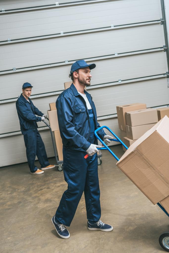 two movers in uniform transporting cardboard boxes with hand trucks in warehouse - Photo, Image