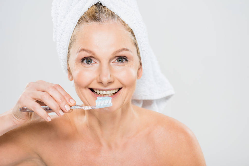 schöne und lächelnde reife Frau in Handtuch Zähne putzen isoliert auf grau  - Foto, Bild