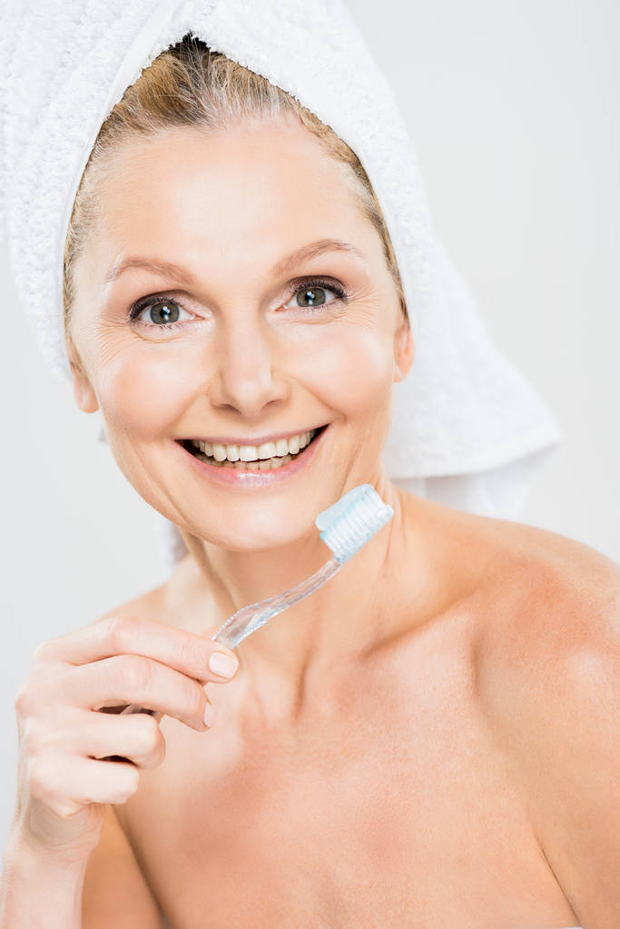 beautiful and smiling mature woman in towel brushing teeth isolated on grey  - Photo, Image