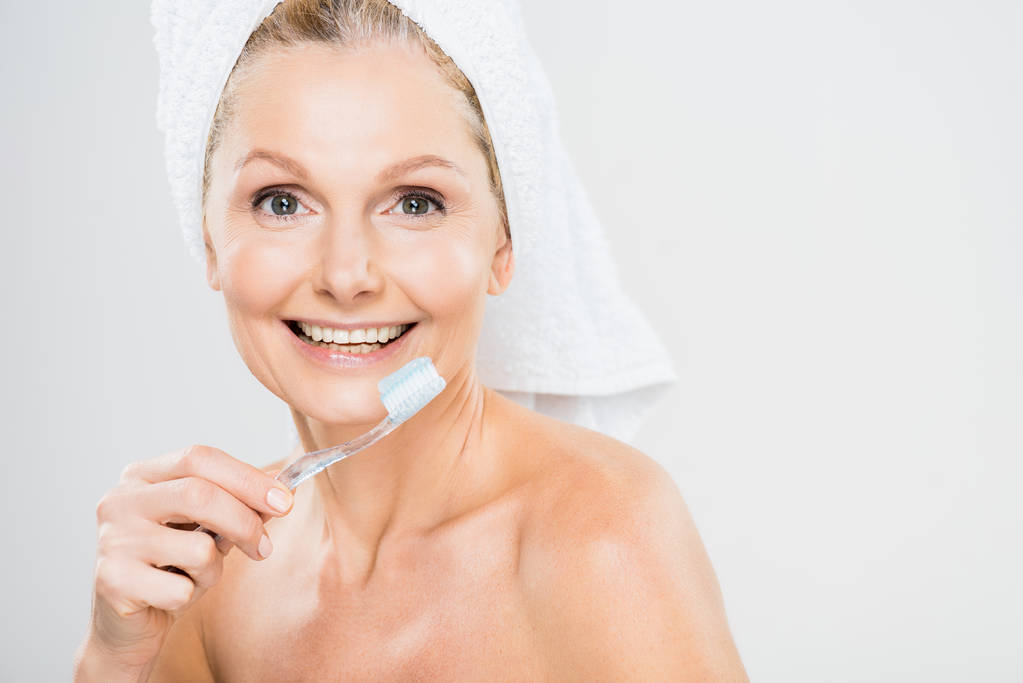 aantrekkelijk en lachende volwassen vrouw in handdoek tandenpoetsen op grijze achtergrond  - Foto, afbeelding