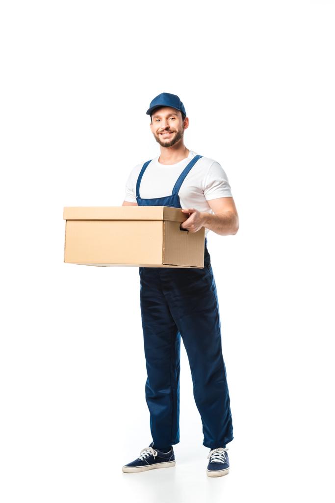 handsome mover in uniform carrying cardboard box, smiling and looking at camera isolated on white - Photo, Image