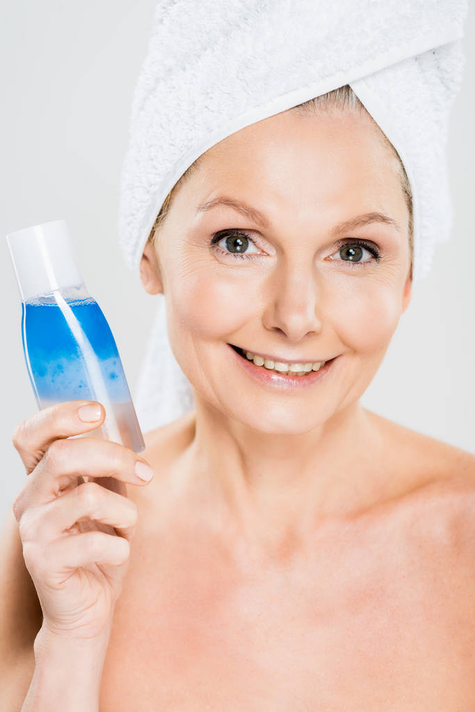 attractive and smiling mature woman in towel holding bottle with micellar water isolated on grey - Photo, Image