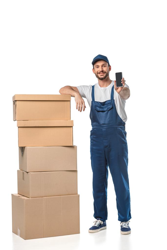 handsome smiling mover with cardboard boxes presenting smartphone with blank screen isolated on white - Photo, Image