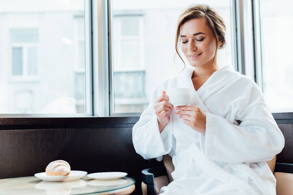 attraktive brünette Frau im Bademantel, die lächelt und auf eine Tasse Kaffee blickt - Foto, Bild