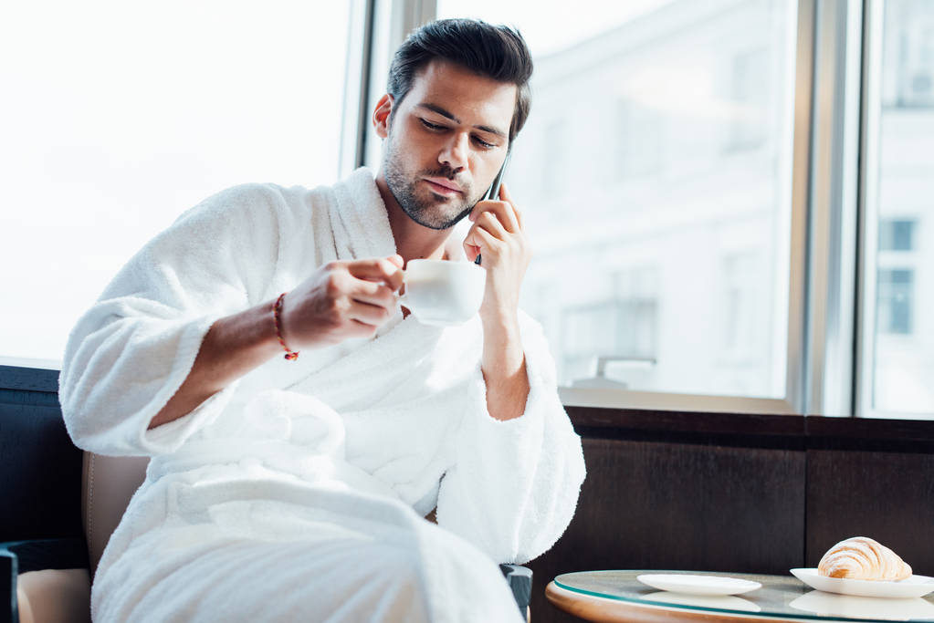  bearded man in white bathrobe holding cup and talking on smartphone  - Photo, Image