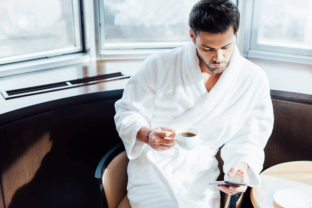 vista aérea del hombre barbudo guapo en albornoz blanco sosteniendo teléfono inteligente y taza con café
 - Foto, Imagen