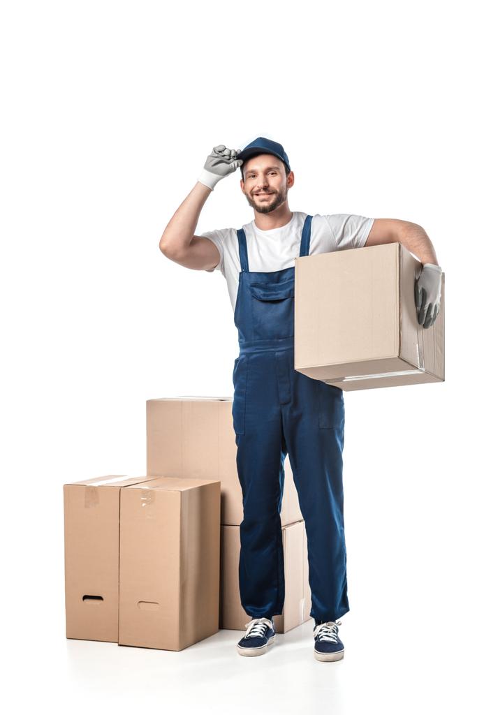 handsome mover in uniform adjusting hat and looking at camera while carrying cardboard box isolated on white - Photo, Image