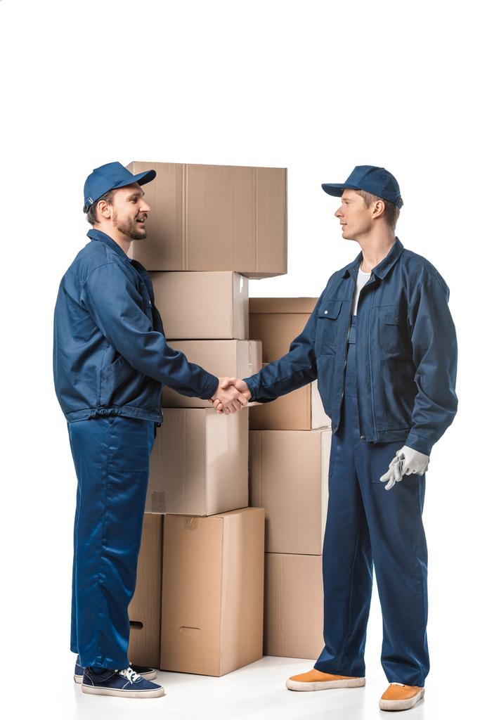 two movers in uniform looking at each other and shaking hands near cardboard boxes on white - Photo, Image