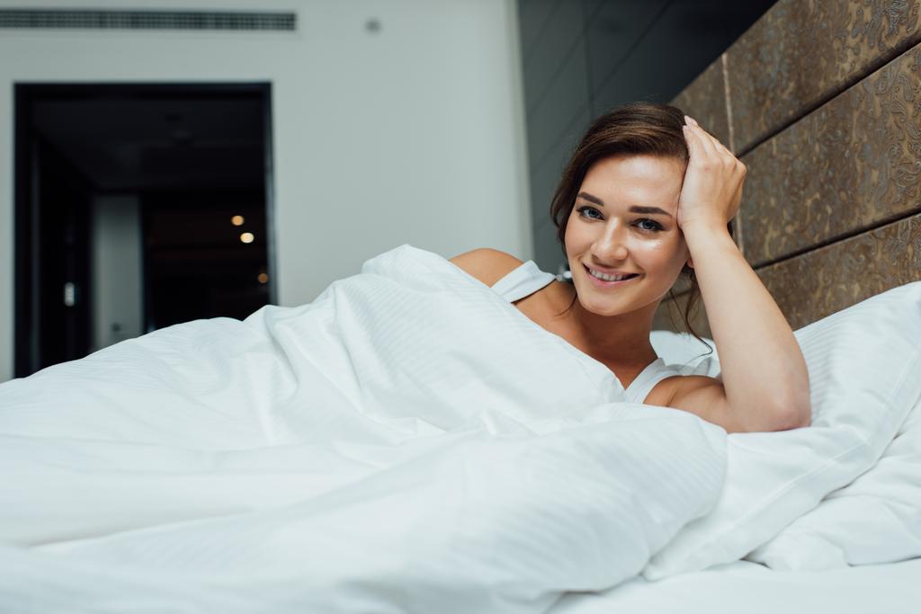 femme brune joyeuse souriant tout en étant couché sur des oreillers sous la couverture au lit
  - Photo, image