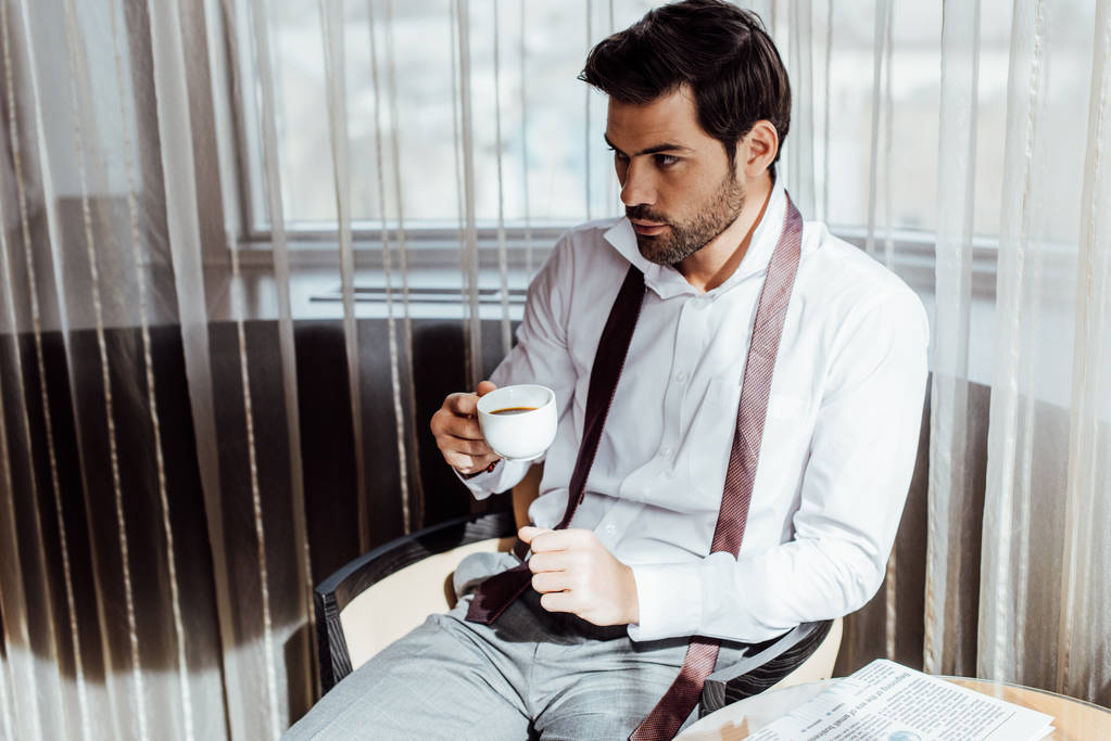 όμορφος άντρας με κοστούμι κάθεται κοντά στο τραπεζάκι με εφημερίδα και κρατώντας το φλιτζάνι του καφέ - Φωτογραφία, εικόνα