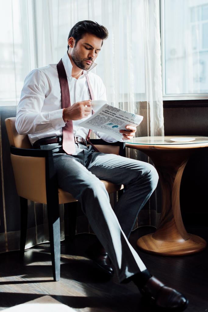 handsome man in suit sitting near coffee table while reading newspaper and holding cup of coffee - Photo, Image