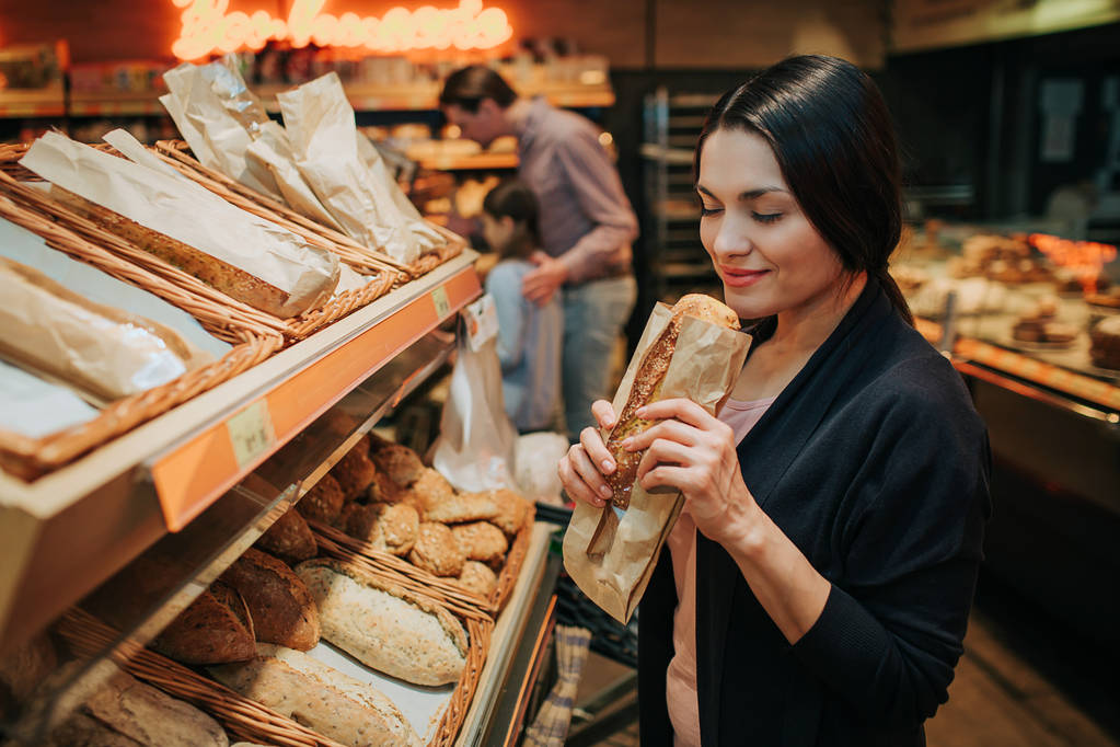 Fiatal szülők és a lánya az élelmiszerboltban. Vidám nő illata kenyeret ő kezében. Apa állni a lánya és a carry élelmiszerbolt targonca. - Fotó, kép