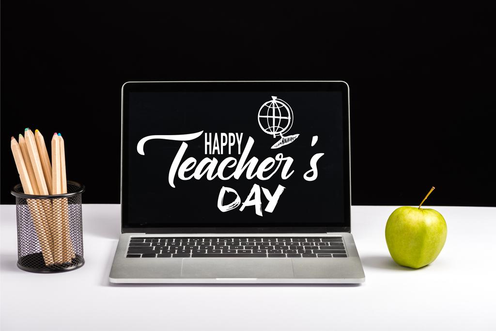 čerstvé zelené jablko, barevné tužky a laptop na stole s happy učitelé den nápisy a koule na obrazovce izolované na černém - Fotografie, Obrázek