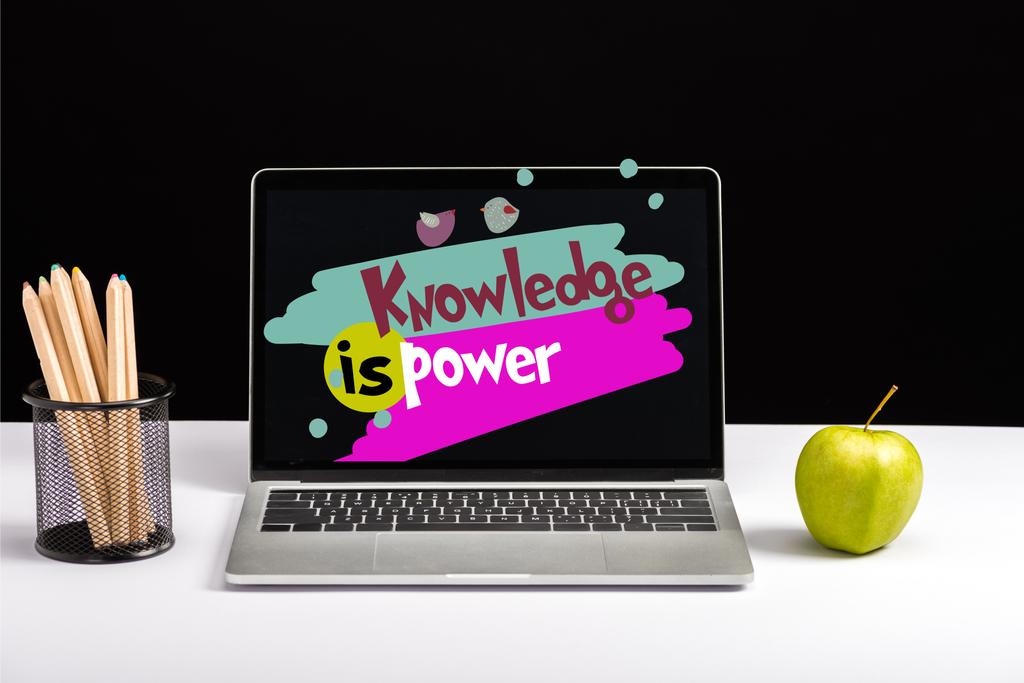 fresh green apple, color pencils and laptop on table with knowledge is power lettering on screen isolated on black - Photo, Image