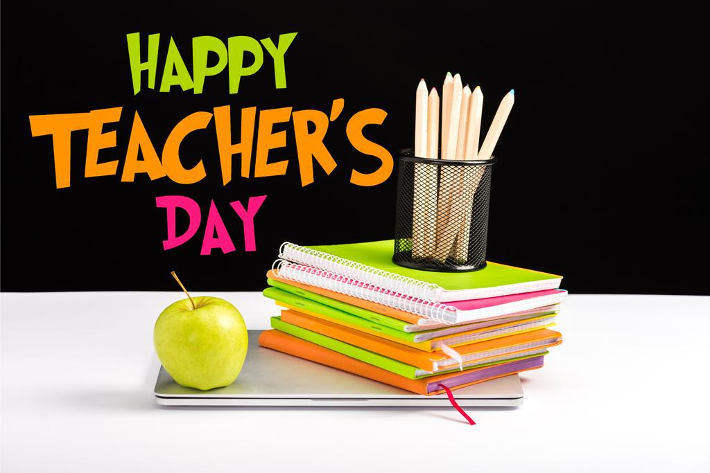 geschlossener Laptop, grüner Apfel, Notizbücher und Buntstifte auf dem Schreibtisch mit glücklichen Lehrern Tagesschrift auf schwarz  - Foto, Bild