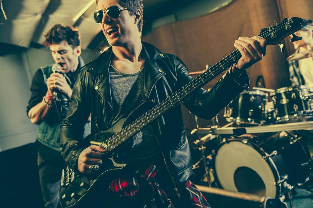 Selektivní fokus veselá kytarista v sluneční brýle hrát elektrická kytara poblíž rocková kapela  - Fotografie, Obrázek