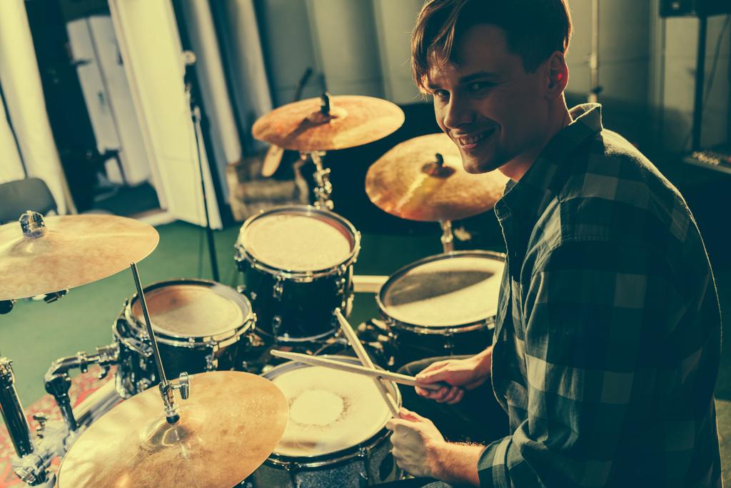 alegre músico guapo sosteniendo palos de tambor cerca de tambores
 - Foto, Imagen
