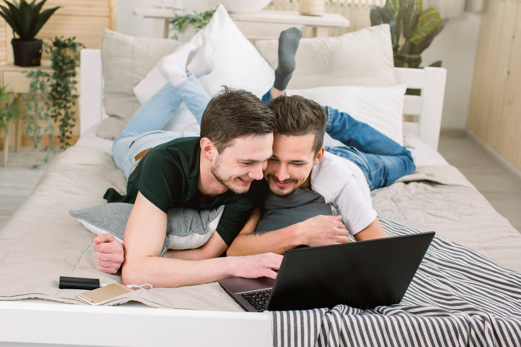 Привлекательный молодой человек опирается на своего парня в постели, наблюдает за ноутбуком. Пара геев лежит на кровати и использует ноутбук дома
 - Фото, изображение
