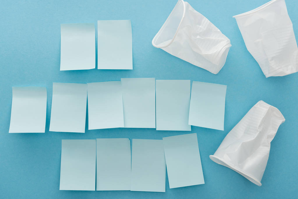 Draufsicht auf weiße Plastik und zerknüllte Tassen und leere Haftnotizen auf blauem Hintergrund  - Foto, Bild