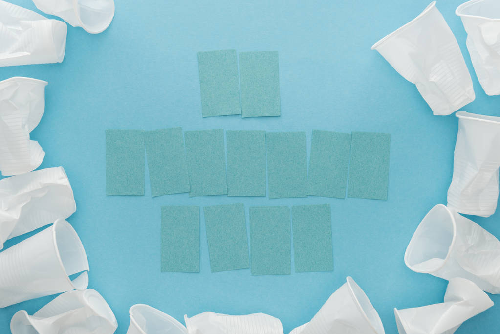 bovenaanzicht van witte plastic bekers en lege kleverige nota's met kopie ruimte op blauwe achtergrond  - Foto, afbeelding