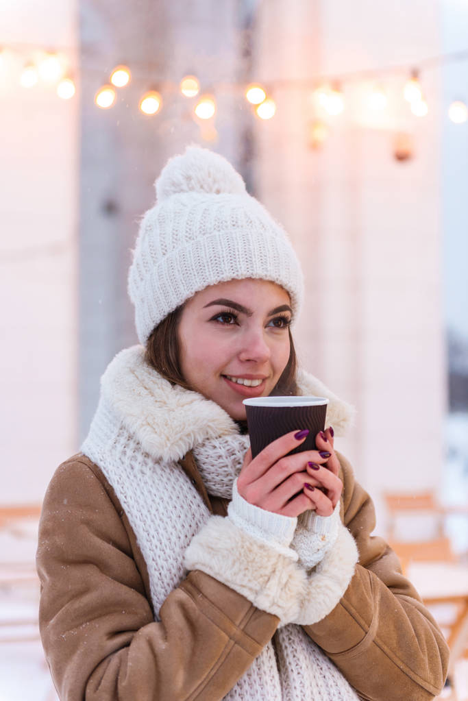 Νεαρή γυναίκα με καπέλο και κασκόλ περπάτημα σε εξωτερικούς χώρους τον χειμώνα χιόνι πίνοντας καφέ. - Φωτογραφία, εικόνα