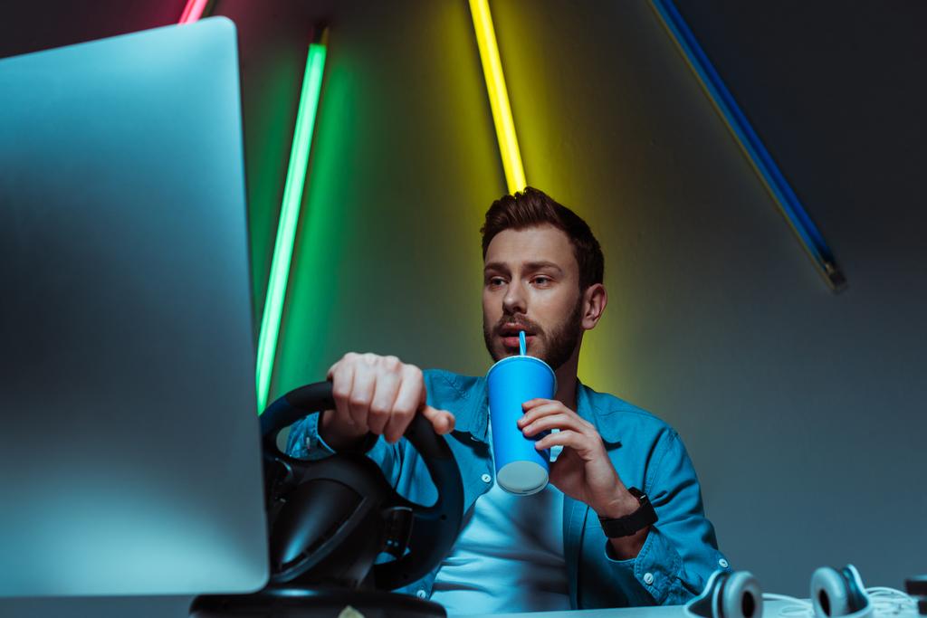 komea ja hyvännäköinen mies juo limua ja pelaa videopeliä ohjauspyörällä
 - Valokuva, kuva