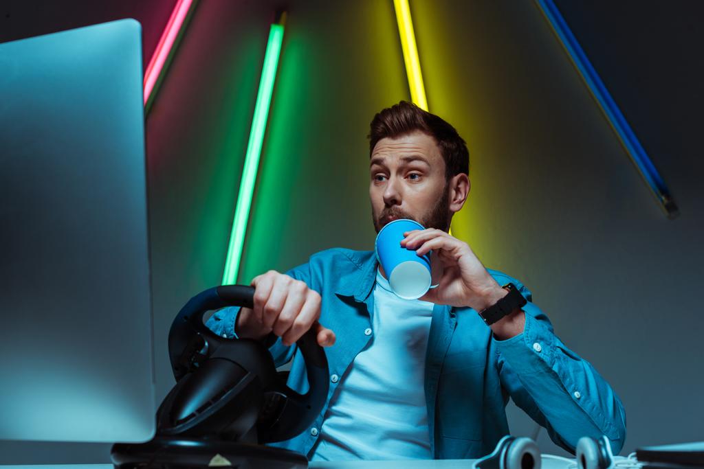 όμορφος και εμφανίσιμος άνθρωπος πίνοντας σόδα και παίζοντας βιντεοπαιχνίδι με το τιμόνι - Φωτογραφία, εικόνα
