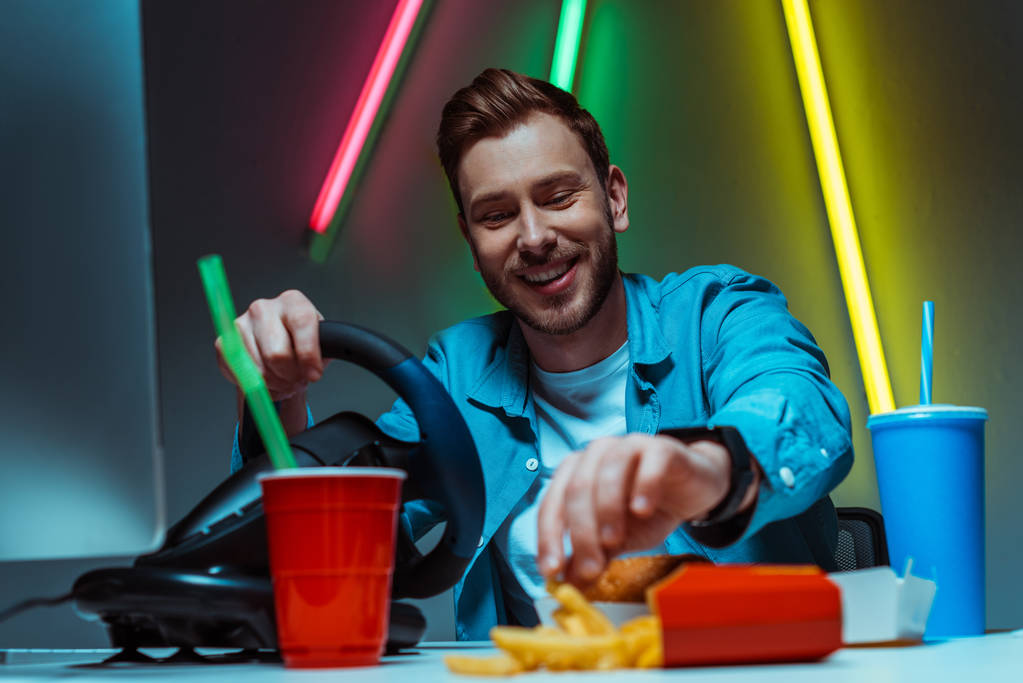selective focus of handsome and smiling man holding steering wheel and eating french fries - Photo, Image
