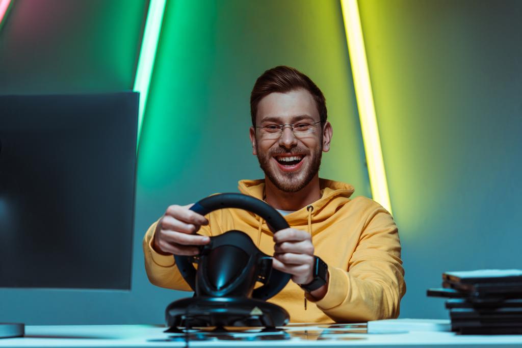 χαμογελαστός, εμφανίσιμος και όμορφος άντρας σε γυαλιά παίζοντας βιντεοπαιχνίδι με το τιμόνι - Φωτογραφία, εικόνα