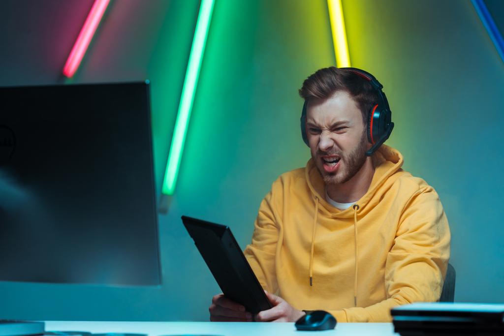 θυμωμένος και όμορφος άνθρωπος στα ακουστικά κρατώντας το πληκτρολόγιο του υπολογιστή και κοιτάζοντας την οθόνη του υπολογιστή  - Φωτογραφία, εικόνα