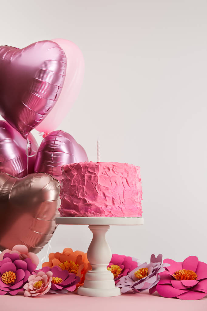 pink birthday cake with candle on cake stand near paper flowers and heart-shaped air balloons on grey - Photo, Image