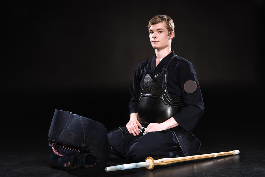 Jonge man in kendo harnas met helm en bamboe zwaard zittend op zwart - Foto, afbeelding