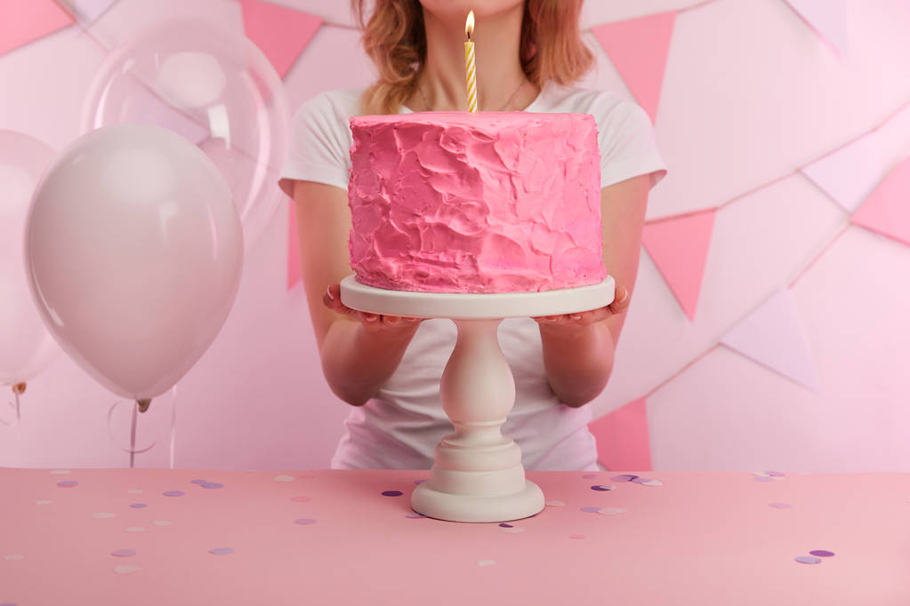 vista recortada de la mujer de pie cerca de sabroso pastel de cumpleaños rosa con vela ardiente en el soporte de la torta
  - Foto, Imagen