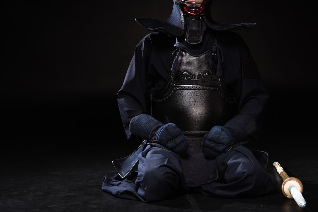 Partial view of kendo fighter in helmet with bamboo sword sitting on black - Photo, Image