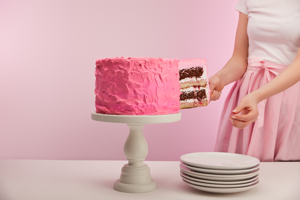 обрезанный вид женщины, принимающей кусок сладкого торта на день рождения возле белых тарелок на розовом
 - Фото, изображение