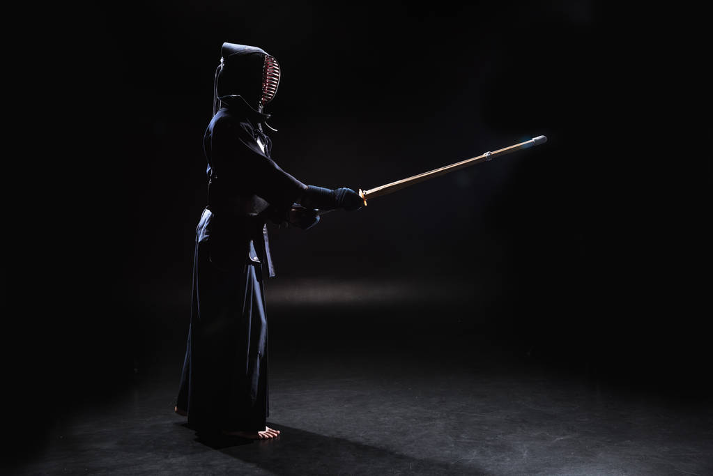 Боковой вид кэндо-бойца в доспехах, практикующегося с бамбуковым мечом на черном
 - Фото, изображение