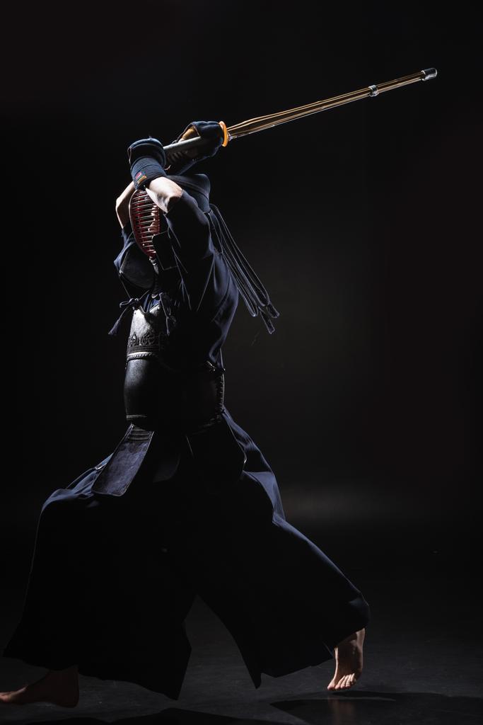 Volle lengte weergave van kendo vechter in harnas oefenen met bamboe zwaard op zwart - Foto, afbeelding