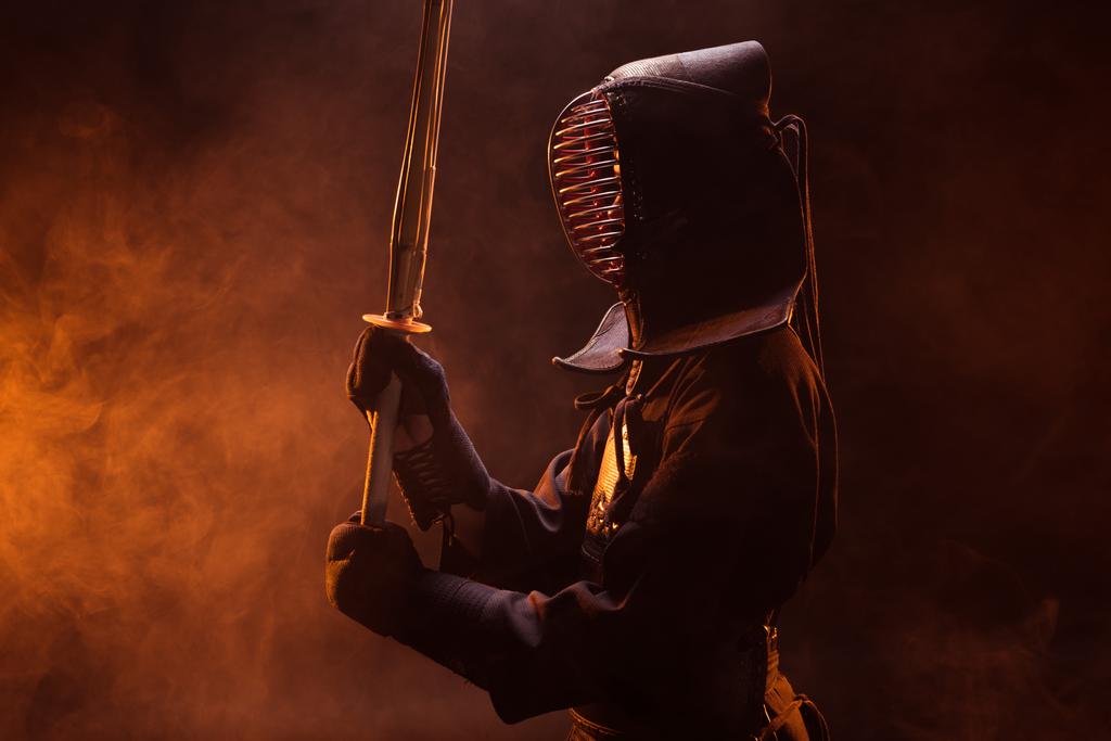 Zijaanzicht van kendo vechter in harnas houden van bamboe zwaard in rook - Foto, afbeelding