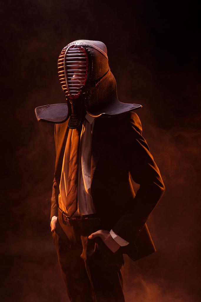 Уверенный боец кендо в формальной одежде и шлеме, стоящий с руками в карманах в темноте
 - Фото, изображение