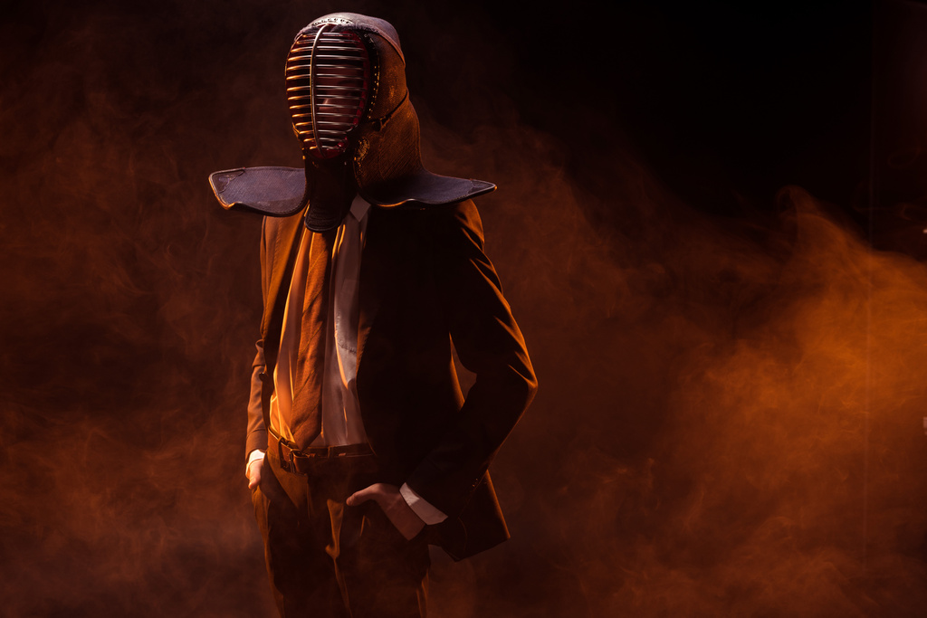 selbstbewusster Mann in formeller Kleidung und Kendo-Helm mit Händen in Taschen auf dunklem - Foto, Bild