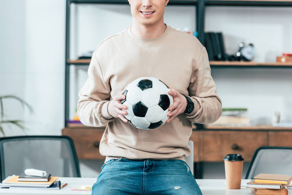 Vista recortada de un joven sonriente sentado en la mesa y sosteniendo la pelota de fútbol en el lugar de trabajo
 - Foto, imagen